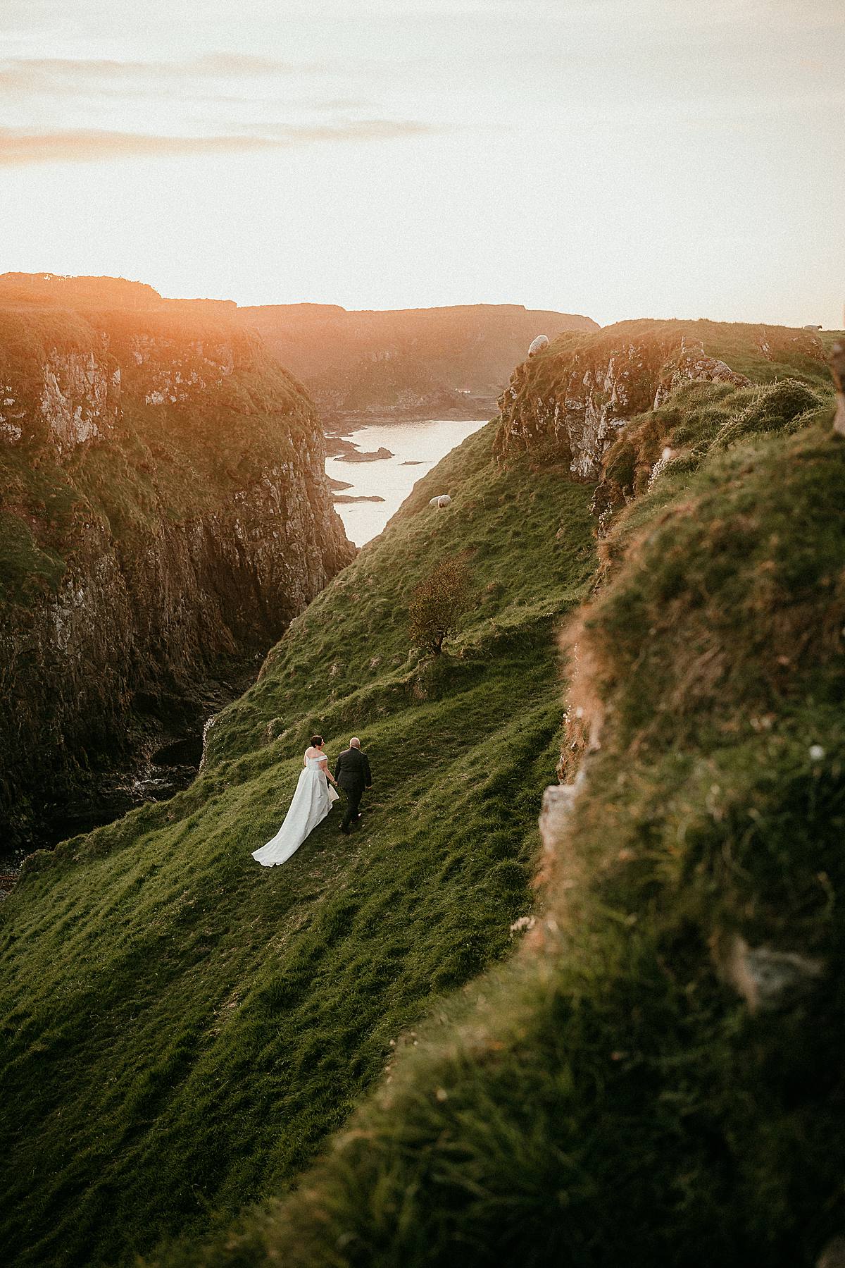 a bride and groom hiking up the grassy side of a cliff at sunset in Northern Ireland on their elopement day.