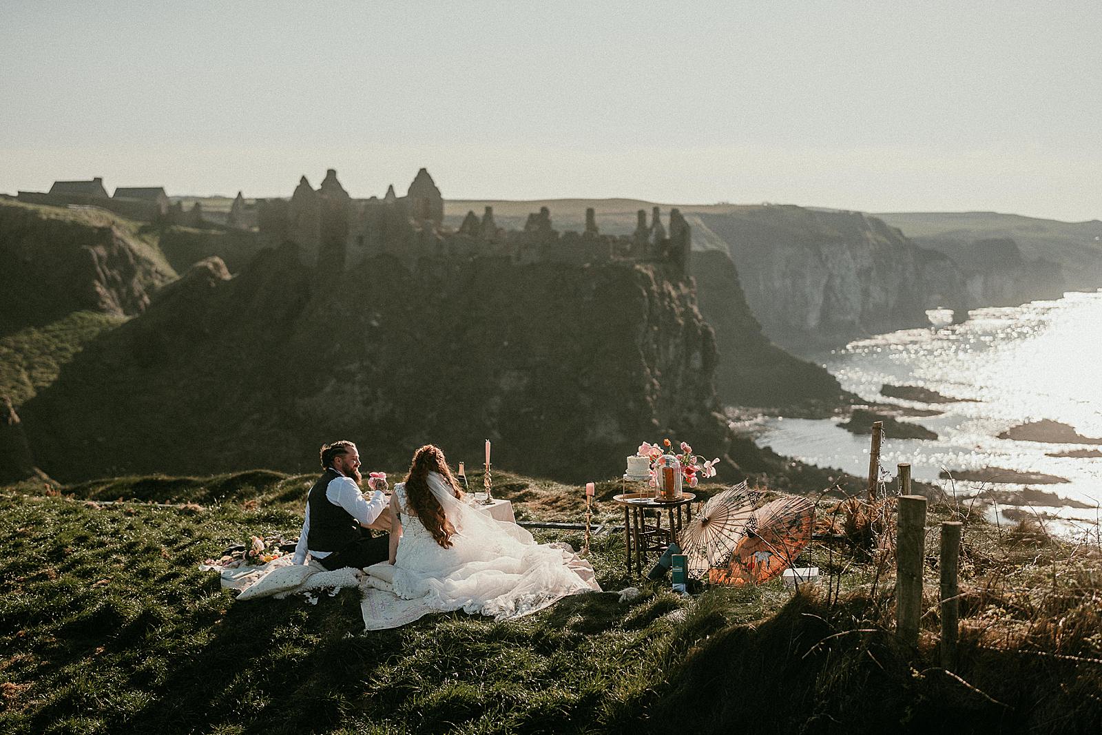 a bride and groom enjoying a luxury picnic after their elopement ceremony overlooking Dunluce Castle on a sunny afternoon.