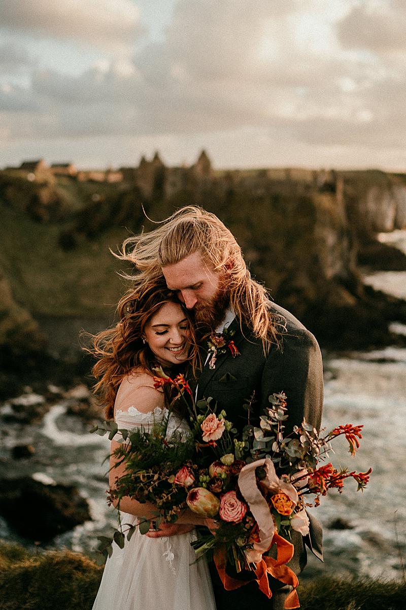 A bride and groom embracing on a cliffside in Northern Ireland with Dunluce Castle in the background, showing wind-swept hair and authentic emotion during an adventure elopement.