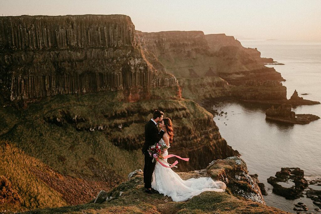 A groom kissing his bride on the forehead stood on a clifftop overlooking the North Atlantic Ocean. The wind is blowing her hair and the sunlight looks like golden hour on their Ireland Elopement day.