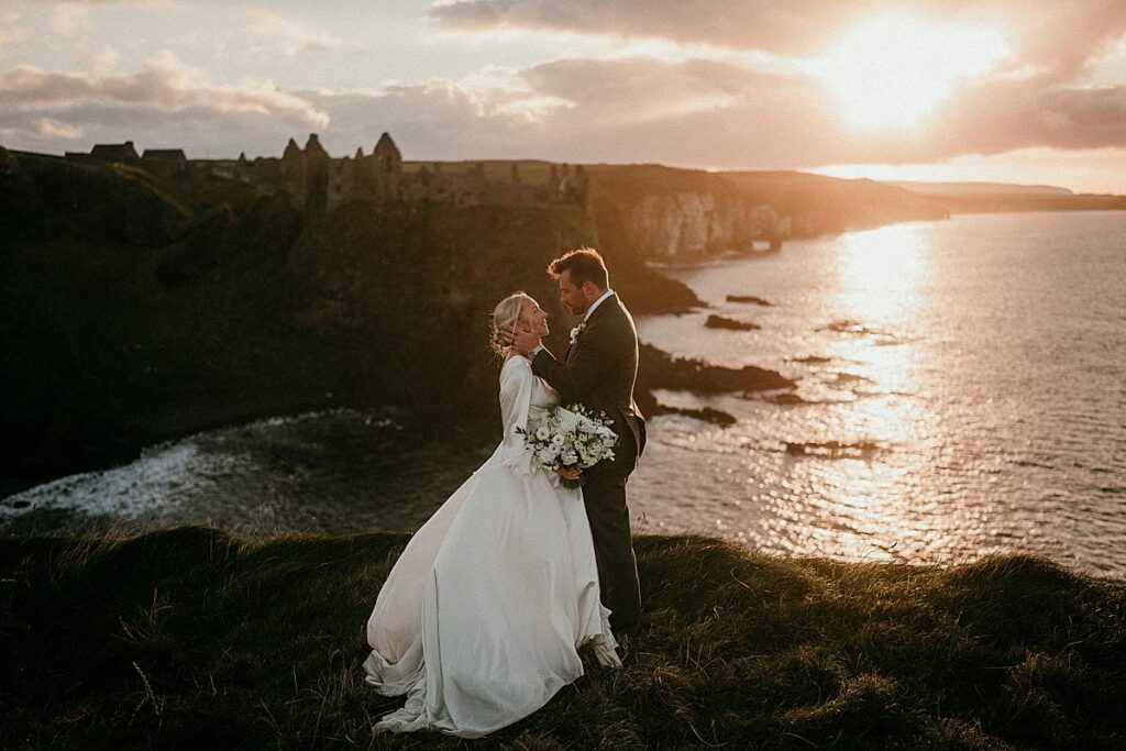 A bride & groom standing on the edge of a cliff top over looking the North Atlantic & Dunluce Castle as the sun is setting causing the sky to glow with golden hour hues.