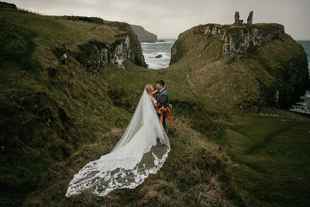 A bride & Groom embracing on the vow renewal day in Ireland. The bride has a huge detailed veil and Dunseverick Castle is in the background.