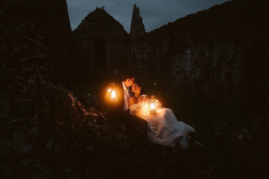A bride & groom in the ruins of an Irish abbey at dusk, they are holding lanterns and it looks very romantic.
