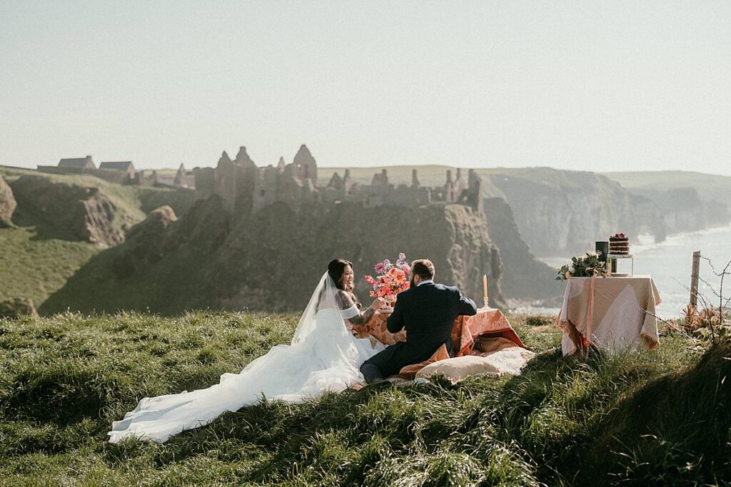 A bride and groom sat having a picnic on the cliff top overlooking Dunluce Castle on their elopement day in Northern Ireland.