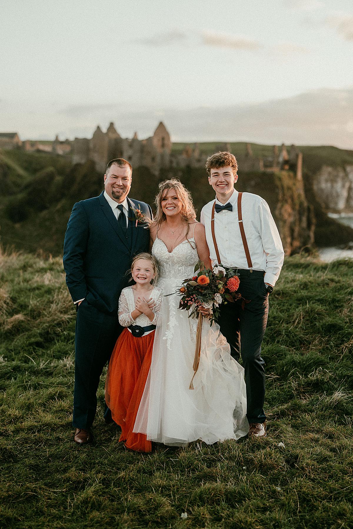 a family group photo of a mum and dad and their teenage son and 6 year old daughter. It is the parents wedding day so they are wearing a wedding dress and a suit. Dunluce Castle is in the background.