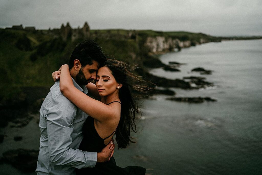 A romantic couple embracing on a cliffside overlooking the ocean at Dunluce Castle, captured by an Ireland couples photographer.