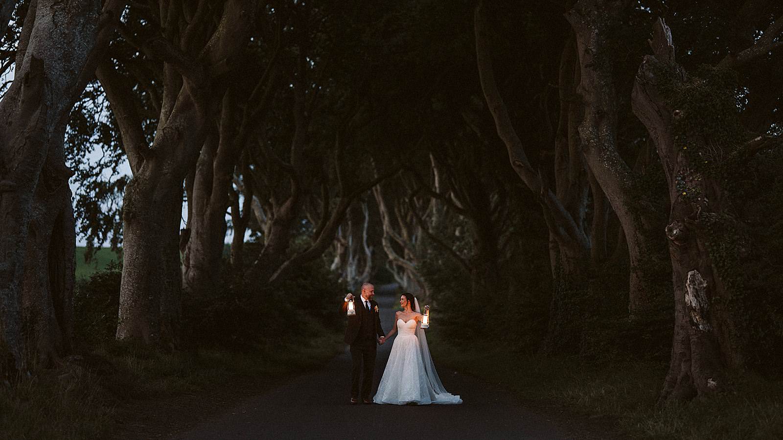 A bride and groom walking hand in hand holding lanterns as they explore the dark Hedges in Northern Ireland on their elopement day.